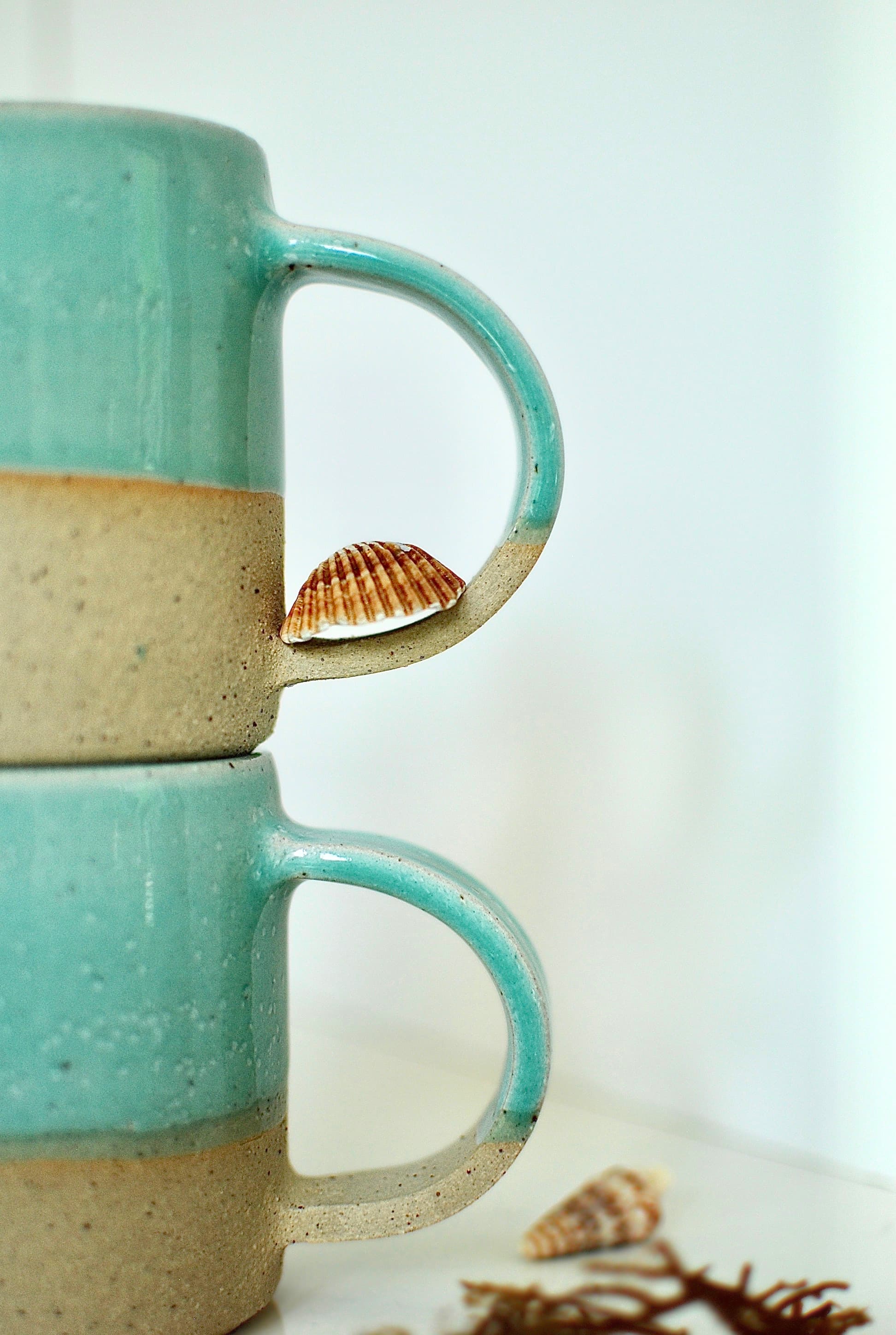 Two handmade stoneware mugs stacked, turquoise glaze over speckled natural clay, coastal styling with seashell β Studio Lippi Ceramics