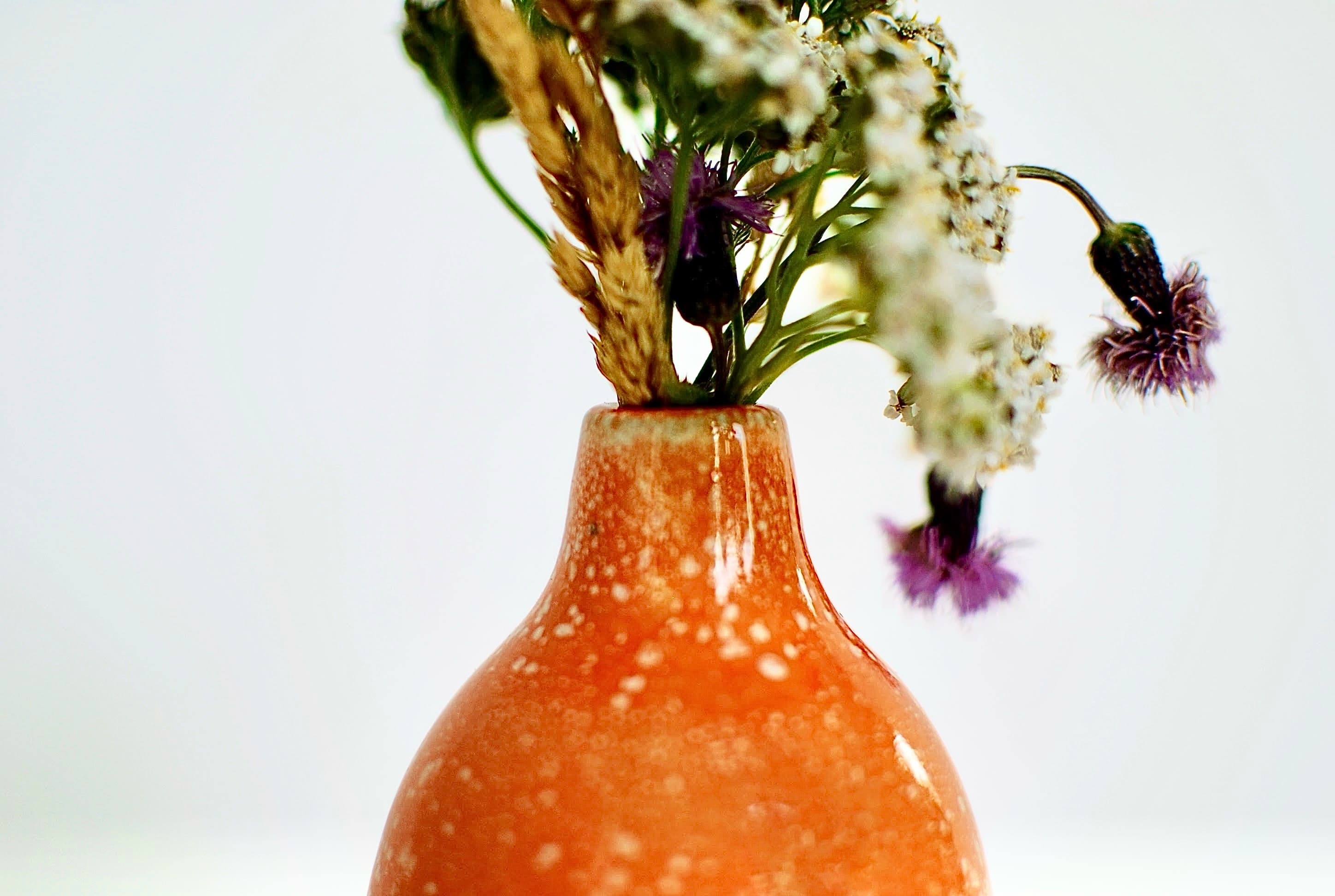 Close-up of the neck of a handmade orange ceramic vase, showing the curved shape and vivid layered orange and white glaze with a speckled finish.