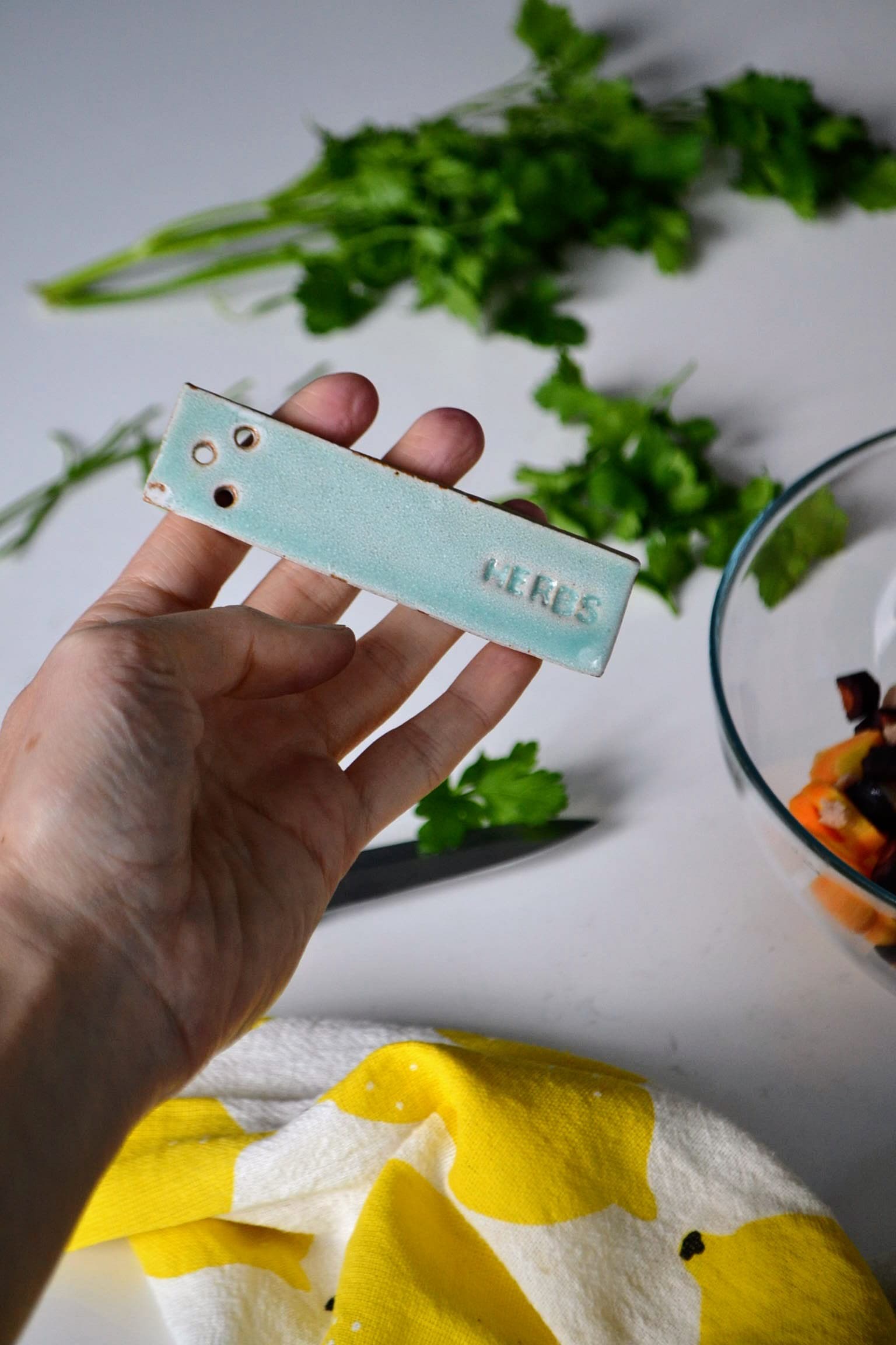 Handmade turquoise ceramic herb stripper held over fresh herbs, showing three holes for stripping herb stems