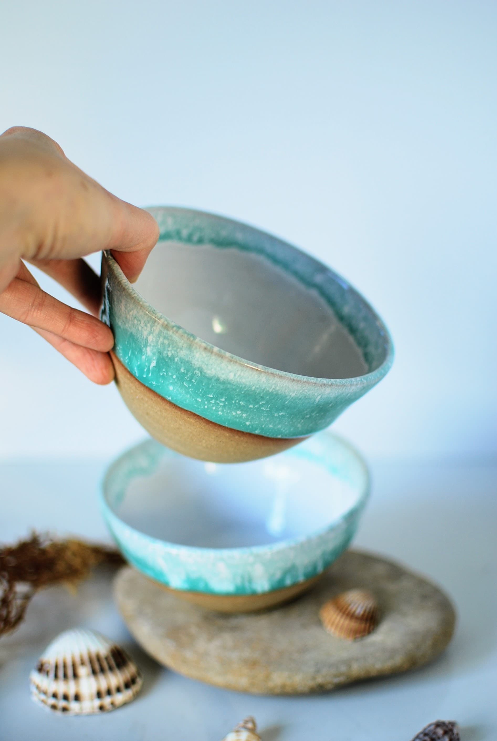Handmade turquoise-glazed stoneware bowl held in hand, with a glossy white interior and natural sandy clay base, crafted by Studio Lippi Ceramics in Dublin.
