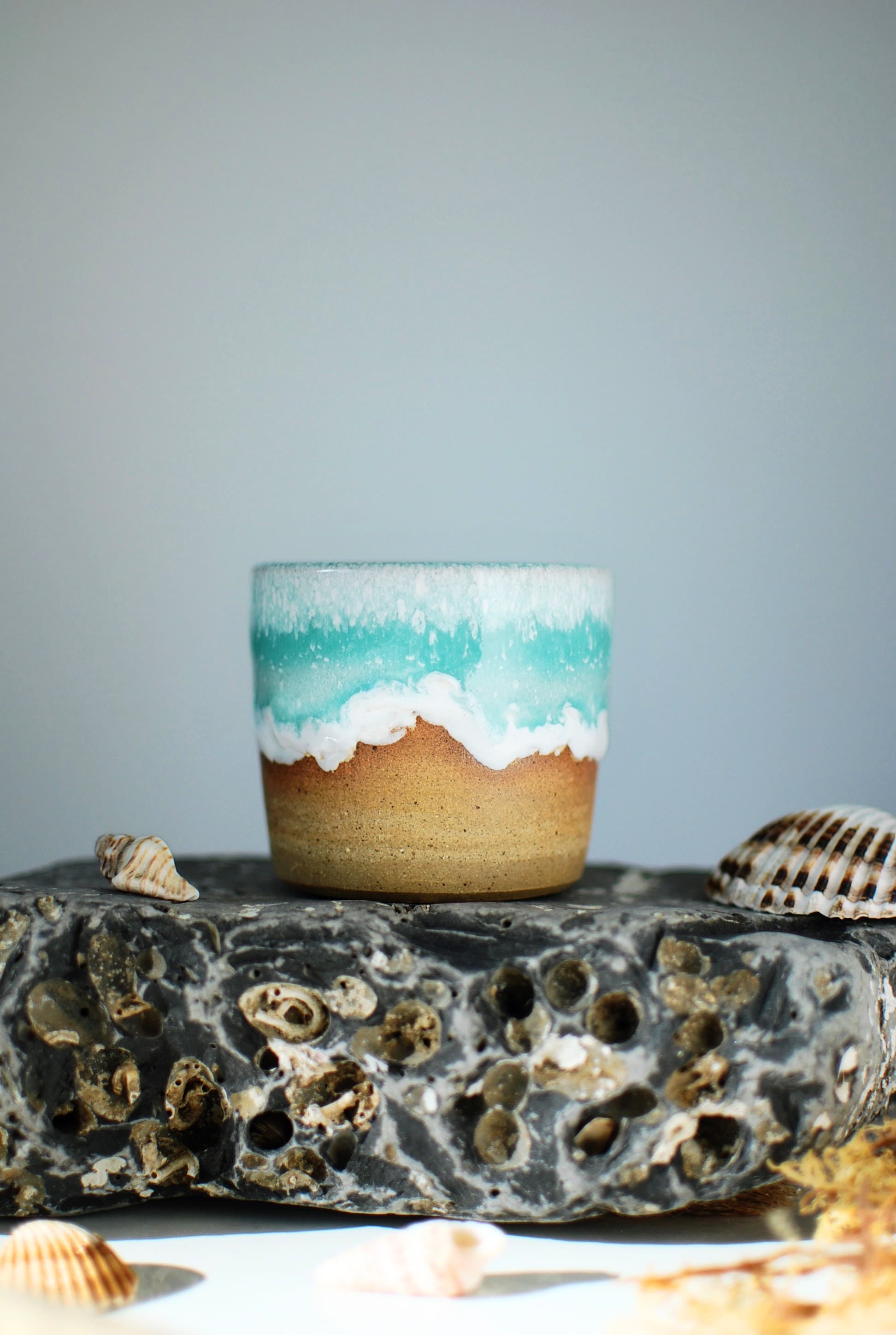 Handmade ceramic espresso cup by Studio Lippi Ceramics, featuring a turquoise and white ocean-inspired glaze on sandy stoneware, photographed on textured rock with seashells.